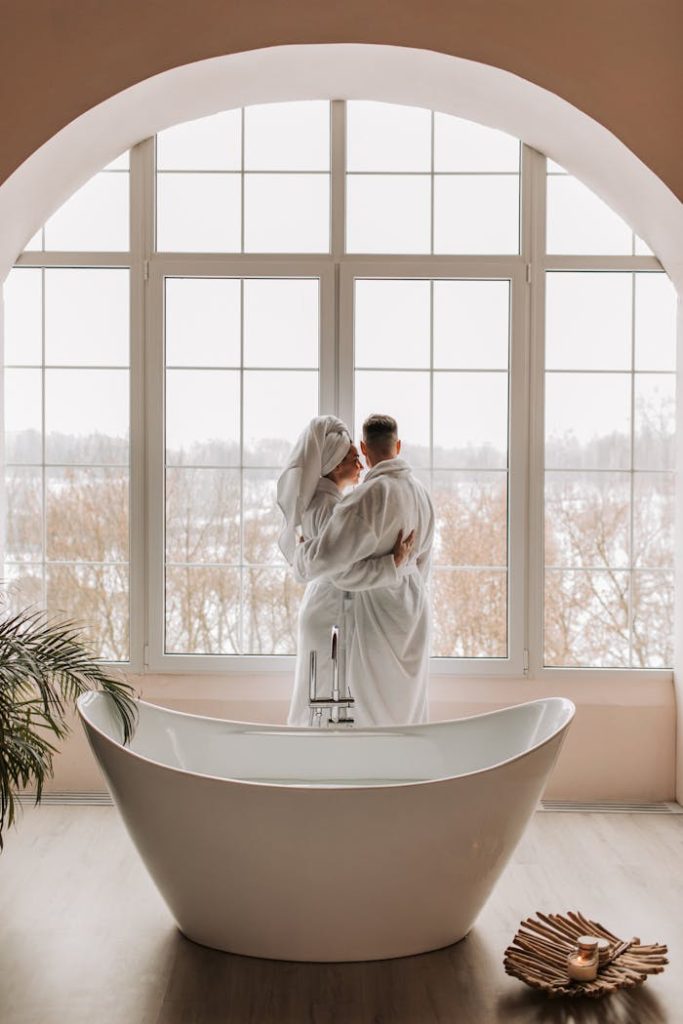 Couple in bathrobes embracing by a window in a luxurious bathroom with a freestanding tub.